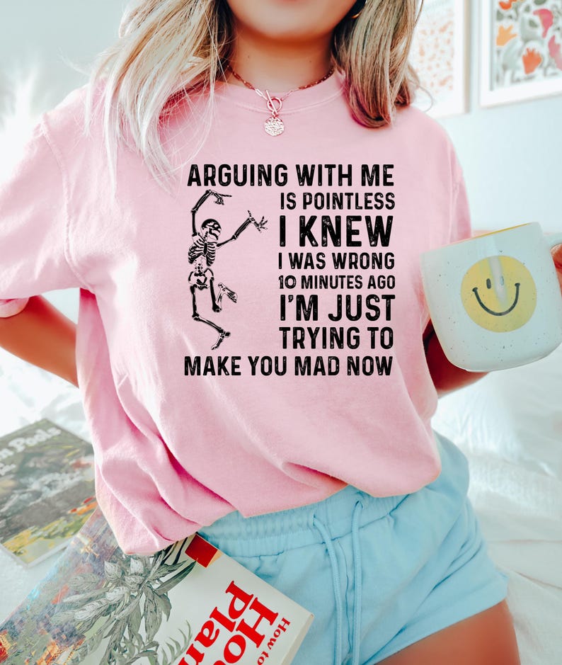 Puede incluir: Sudadera rosa claro con el texto "ARGUING WITH ME IS POINTLESS I KNEW I WAS WRONG 10 MINUTES AGO I'M JUST TRYING TO MAKE YOU MAD NOW" y un gr&aacute;fico de esqueleto. Se sostiene una taza blanca con una carita sonriente amarilla.