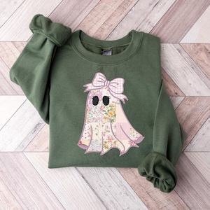 May include: An olive green sweatshirt featuring a ghost applique. The ghost has a floral pattern body, a pink gingham bow, and black eyes. The sleeves are cuffed. The sweatshirt is laying on a light wood surface.