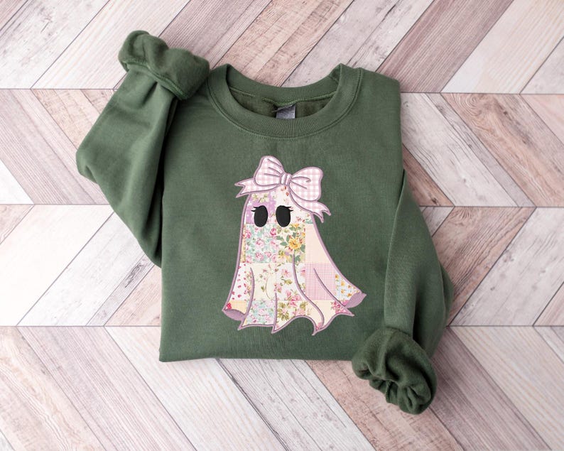 May include: An olive green sweatshirt featuring a ghost applique. The ghost has a floral pattern body, a pink gingham bow, and black eyes. The sleeves are cuffed. The sweatshirt is laying on a light wood surface.