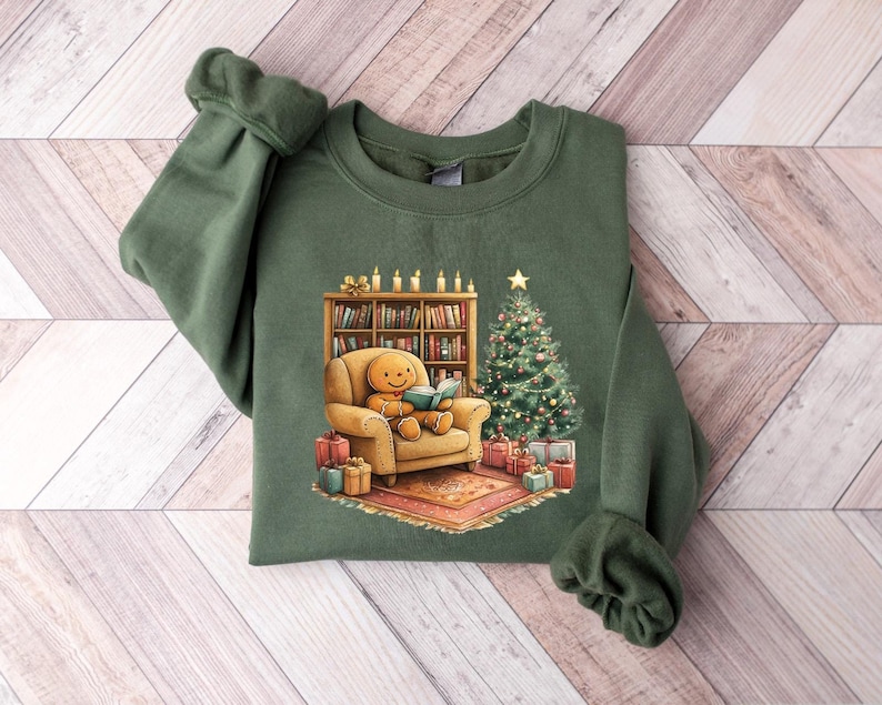May include: A green sweatshirt with a Christmas-themed design featuring a gingerbread person sitting in a chair reading a book in front of a fireplace and a Christmas tree.