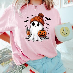 May include: A pink t-shirt with a cartoon ghost wearing a brown and orange knit hat and holding a pumpkin and a beverage. The ghost is surrounded by bats and stars.