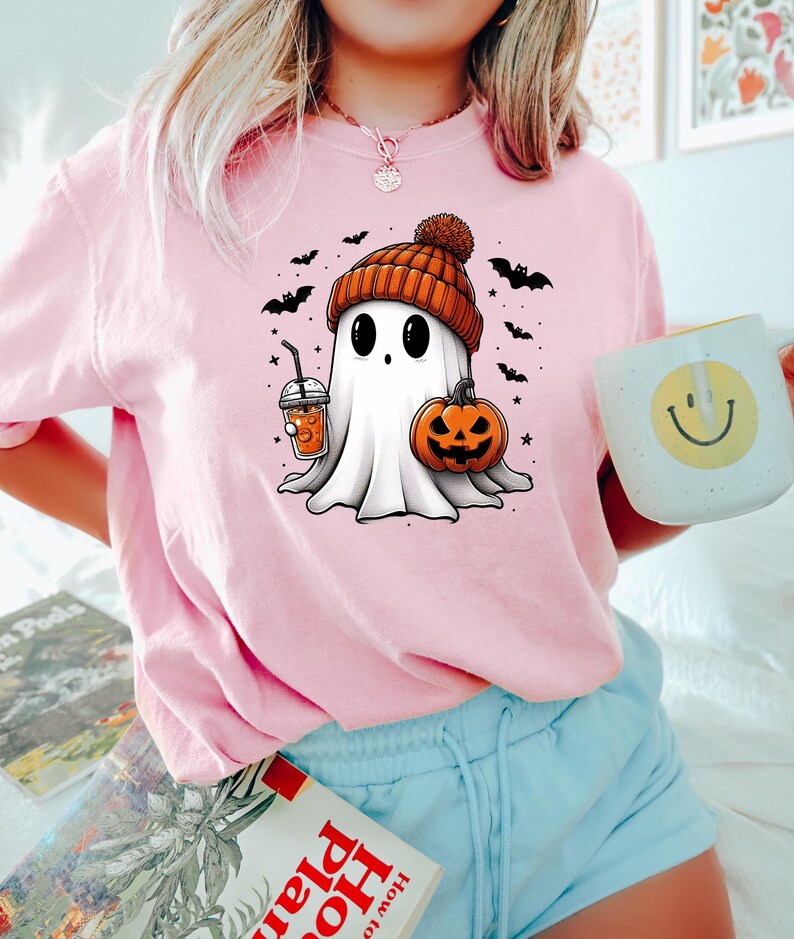May include: A pink t-shirt with a cartoon ghost wearing a brown and orange knit hat and holding a pumpkin and a beverage. The ghost is surrounded by bats and stars.
