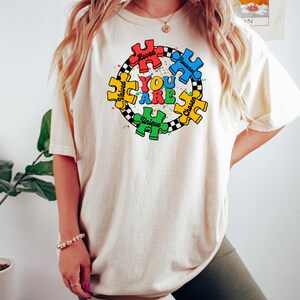 May include: Cream-colored t-shirt with a colorful puzzle piece design. The design features the words "YOU ARE" in blue, surrounded by puzzle pieces in red, blue, green, and yellow, with the words "Loved, Treasured, Chosen, Unique, Valued".