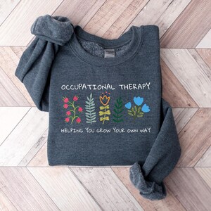 May include: A dark gray sweatshirt with the text "Occupational Therapy" and a graphic of colorful flowers with the text "Helping you grow your own way".