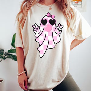 Peut inclure: Un t-shirt blanc avec un fant&ocirc;me &agrave; carreaux rose et blanc portant des lunettes de soleil en forme de c&oelig;ur rose et un n&oelig;ud rose. Le fant&ocirc;me souffle une bulle rose.