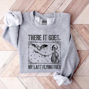 May include: Gray sweatshirt with a black and white graphic of a skeleton holding a bat with the text "There it goes, my last flying f***".