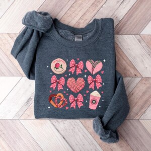 May include: A dark gray sweatshirt with a pink and white graphic print of various Mexican treats, including a heart-shaped pan dulce, a concha, a taco, and a cup of coffee. The text "Mami" is printed on the graphic.