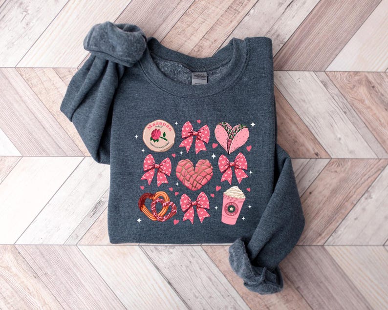 May include: A dark gray sweatshirt with a pink and white graphic print of various Mexican treats, including a heart-shaped pan dulce, a concha, a taco, and a cup of coffee. The text "Mami" is printed on the graphic.