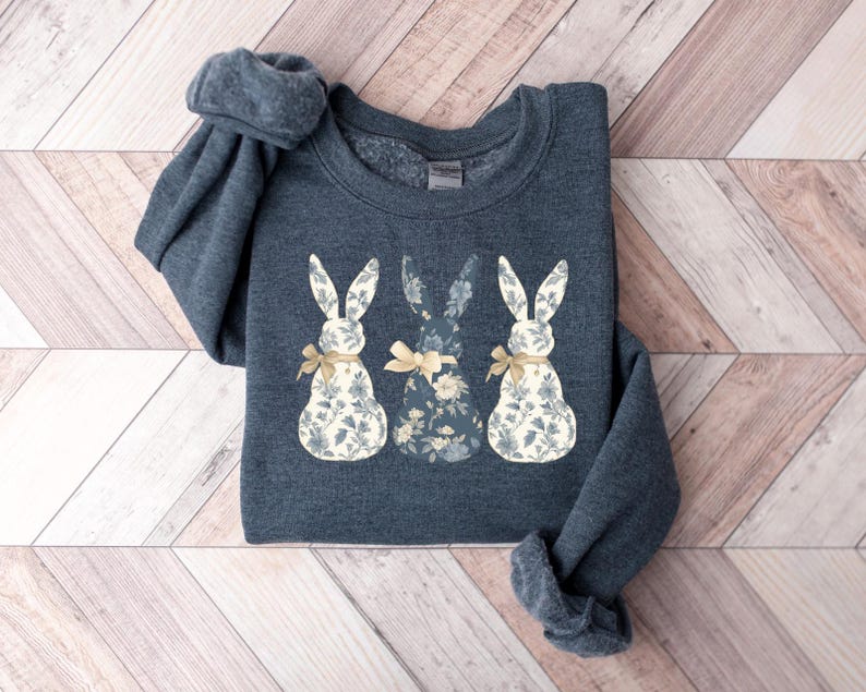 K&ouml;nnte beinhalten: Ein dunkelgrauer Pullover mit einem floralen Muster aus drei Hasen. Die Hasen tragen gelbe Schleifen und haben wei&szlig;e Ohren.