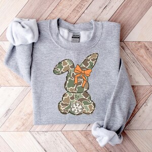 May include: A gray sweatshirt with a camouflage bunny rabbit wearing an orange bow. The bunny is facing away from the viewer.