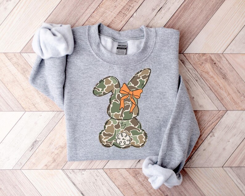 May include: A gray sweatshirt with a camouflage bunny rabbit wearing an orange bow. The bunny is facing away from the viewer.