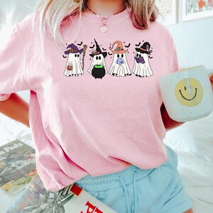 Puede incluir: Camiseta rosa con un gr&aacute;fico de cinco fantasmas con sombreros de bruja. Los fantasmas sostienen una caldera, un libro, una varita m&aacute;gica y una calabaza.