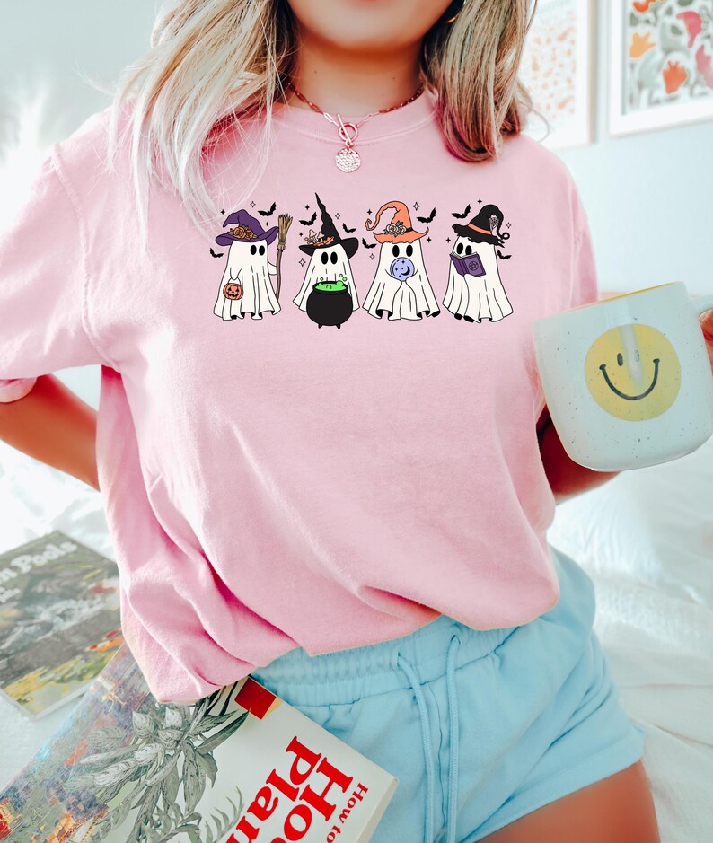 Puede incluir: Camiseta rosa con un gr&aacute;fico de cinco fantasmas con sombreros de bruja. Los fantasmas sostienen una caldera, un libro, una varita m&aacute;gica y una calabaza.