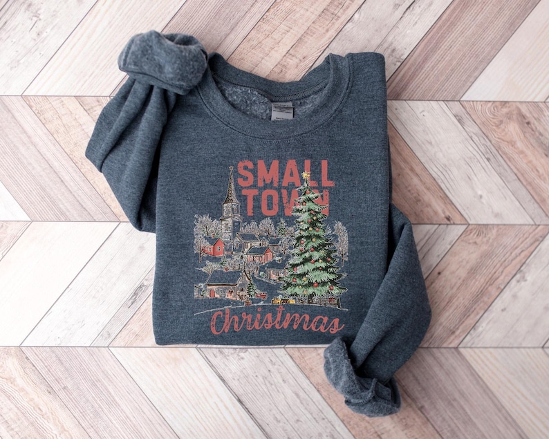 May include: A dark gray sweatshirt with a Christmas-themed design. The design features a small town with a church, houses, and a Christmas tree. The text "Small Town Christmas" is printed in red on the sweatshirt.