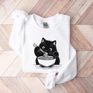 May include: White sweatshirt with a black cat eating ramen noodles from a bowl. The cat is holding chopsticks in its paws.