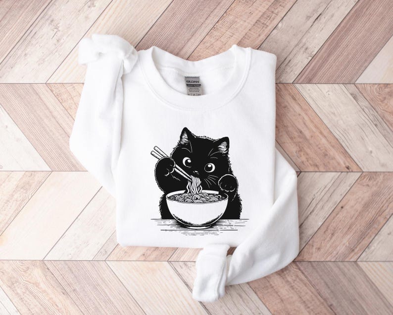 May include: White sweatshirt with a black cat eating ramen noodles from a bowl. The cat is holding chopsticks in its paws.