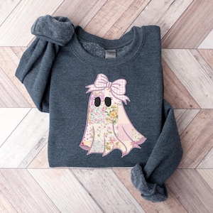 May include: A dark gray sweatshirt featuring a floral ghost design with a pink bow. The ghost has a patchwork design with a floral pattern and black eyes. The sweatshirt is folded on a wooden surface.
