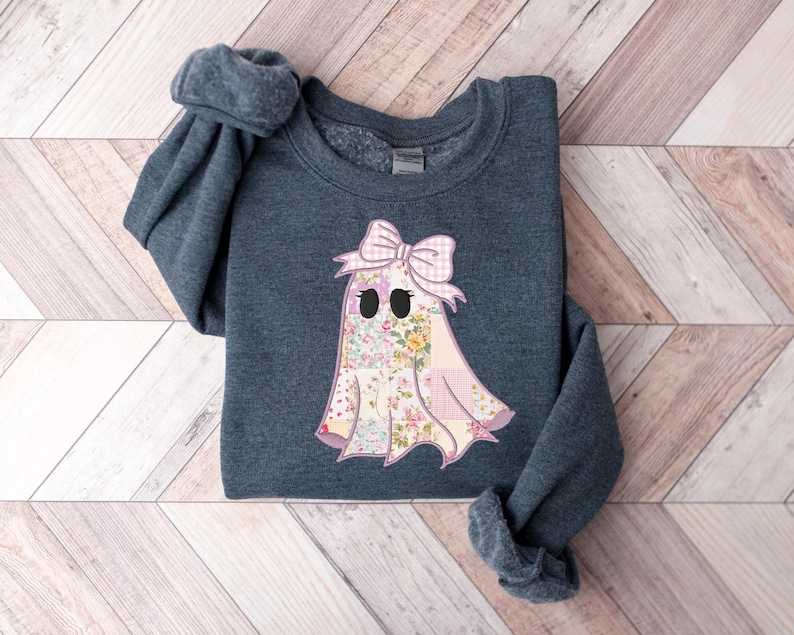 May include: A dark gray sweatshirt featuring a floral ghost design with a pink bow. The ghost has a patchwork design with a floral pattern and black eyes. The sweatshirt is folded on a wooden surface.