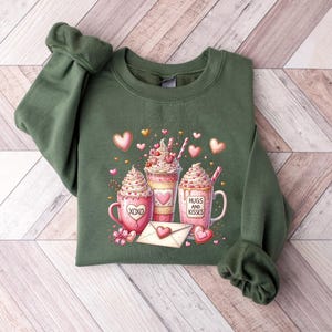 May include: Green sweatshirt with a graphic of three pink and white hot chocolate mugs with whipped cream and sprinkles. The mugs have the text "Hugs and Kisses" and "XOXO" on them. The graphic is surrounded by pink hearts and gold glitter.