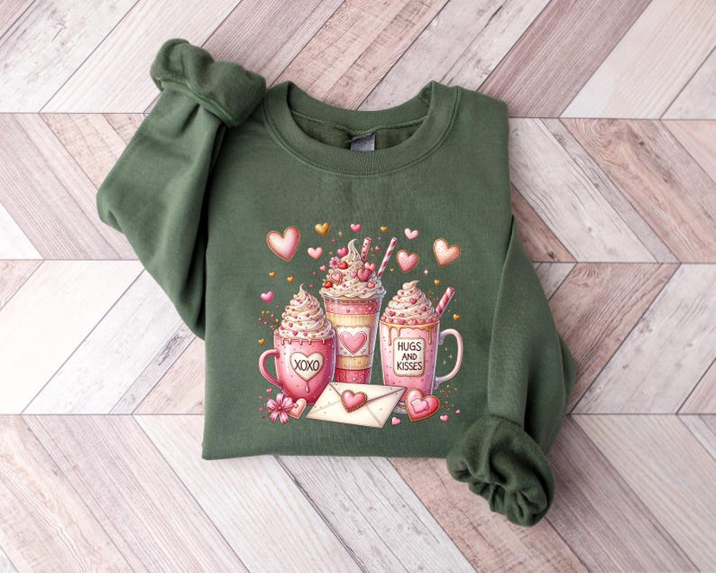 May include: Green sweatshirt with a graphic of three pink and white hot chocolate mugs with whipped cream and sprinkles. The mugs have the text "Hugs and Kisses" and "XOXO" on them. The graphic is surrounded by pink hearts and gold glitter.