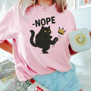 以下が含まれることがあります： 「-NOPE」の文字と、金の王冠を蹴る黒猫のイラストがプリントされたピンク色のスウェットシャツ。前面に猫のイラストが描かれています。黄色のスマイリーフェイスが描かれた白いマグカップが手に持たれています。