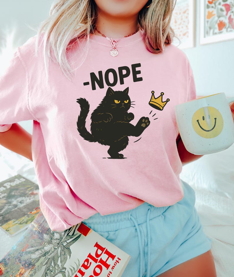 以下が含まれることがあります： 「-NOPE」の文字と、金の王冠を蹴る黒猫のイラストがプリントされたピンク色のスウェットシャツ。前面に猫のイラストが描かれています。黄色のスマイリーフェイスが描かれた白いマグカップが手に持たれています。