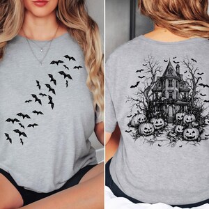 Op de afbeelding: Een grijs T-shirt met een zwarte afbeelding van vliegende vleermuizen aan de voorkant en een spookhuis met pompoenen aan de achterkant.