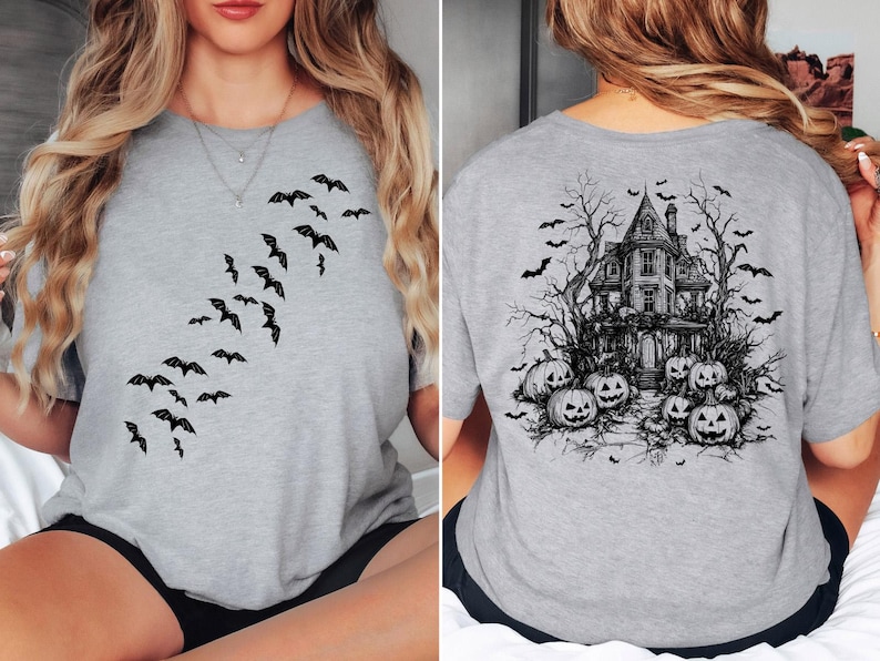 Op de afbeelding: Een grijs T-shirt met een zwarte afbeelding van vliegende vleermuizen aan de voorkant en een spookhuis met pompoenen aan de achterkant.