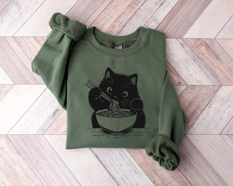 May include: A green sweatshirt with a black cat eating ramen noodles from a bowl. The cat is holding chopsticks in its paws.