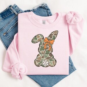 May include: Pink sweatshirt with a camouflage bunny wearing an orange bow. The bunny is sitting down with its back to the viewer.