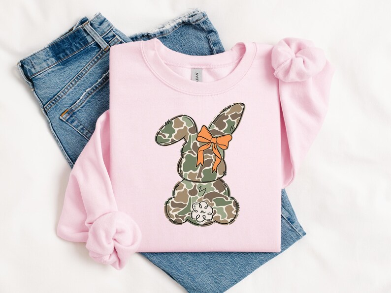 May include: Pink sweatshirt with a camouflage bunny wearing an orange bow. The bunny is sitting down with its back to the viewer.