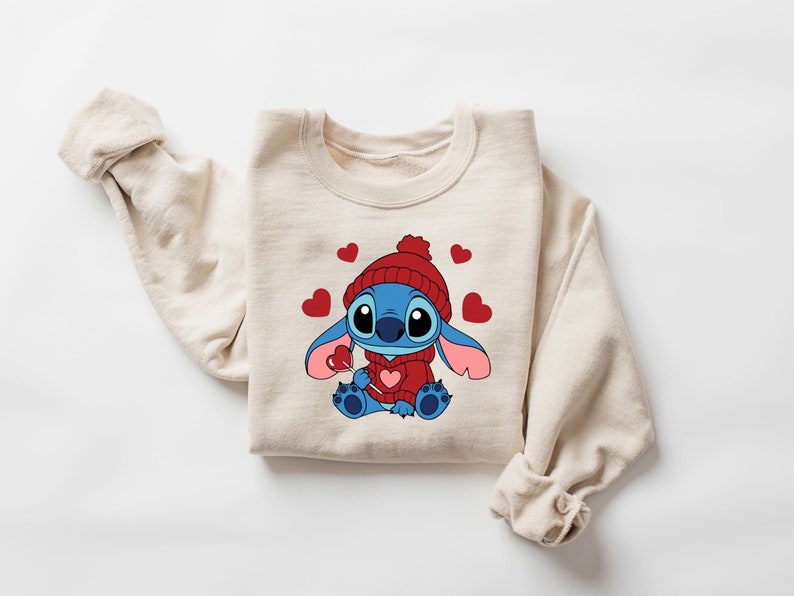 Pu&ograve; includere: Felpa beige con un personaggio dei cartoni animati, Stitch, che indossa un cappello e un maglione rossi, con in mano un lecca-lecca a forma di cuore. Cuori rossi sono sparsi intorno al personaggio. La felpa ha le maniche lunghe.