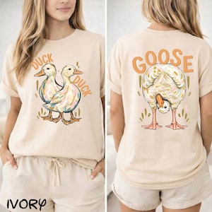 Puede incluir: Camiseta de color marfil con un dise&ntilde;o de pato en la parte delantera y un dise&ntilde;o de ganso en la parte trasera. La parte delantera muestra dos patos con la palabra "DUCK". La parte trasera muestra un ganso con la palabra "GOOSE" encima. La palabra "IVORY" est&aacute; impresa abajo.