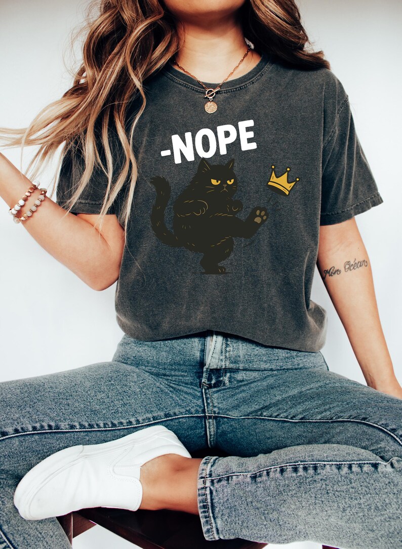 以下が含まれることがあります： 白文字で「-NOPE」と書かれた濃いグレーのTシャツ。黄色い目の黒猫と金の王冠のグラフィックも描かれています。柔らかい素材で作られたカジュアルウェアです。