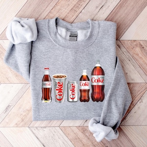 May include: A grey sweatshirt featuring a graphic of four Coca-Cola products: a glass bottle of Diet Coke, a can of Diet Coke, a can of Coke, and a glass bottle of Coke.