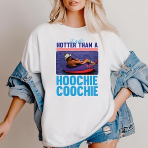 May include: White t-shirt with the text "It gets hotter than a Hoochie Coochie" in blue and a picture of a person floating on a tube in the water. The shirt has a crew neck and short sleeves.