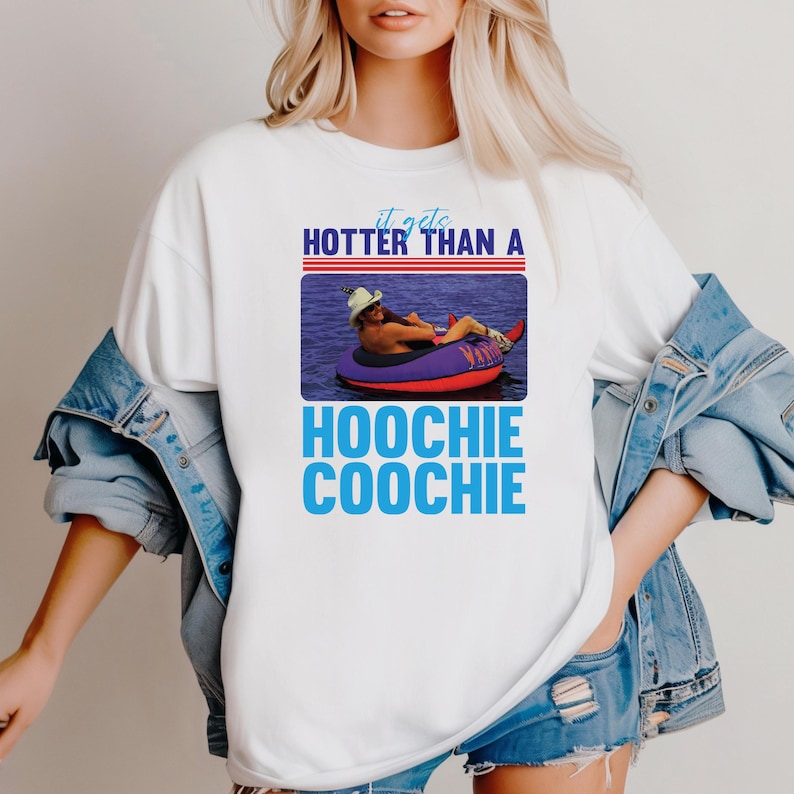 May include: White t-shirt with the text "It gets hotter than a Hoochie Coochie" in blue and a picture of a person floating on a tube in the water. The shirt has a crew neck and short sleeves.