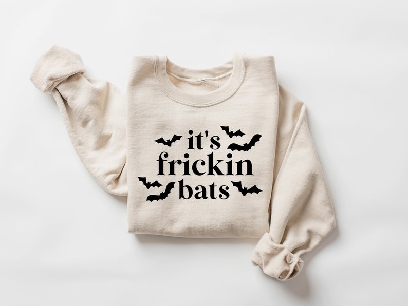 K&ouml;nnte beinhalten: Ein beiger Pullover mit dem Aufdruck "it's frickin bats" in Schwarz und einigen schwarzen Fledermaus-Silhouetten um den Text herum.