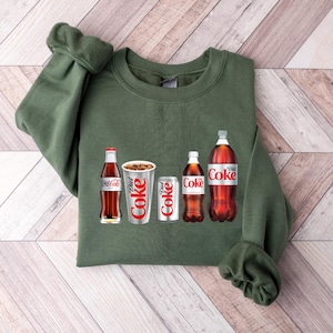 May include: A green sweatshirt featuring a graphic of four Coca-Cola products: a glass bottle of Coca-Cola, a can of Diet Coke, a can of Coca-Cola, and a glass bottle of Diet Coke.