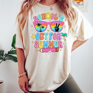Op de afbeelding: Een wit T-shirt met een kleurrijk grafisch ontwerp met de tekst "School's Out For Summer". De grafiek toont een zonnebril met palmbomen en een vredesteken dat in de glazen wordt weerkaatst, een potlood en sterren en bloemen.