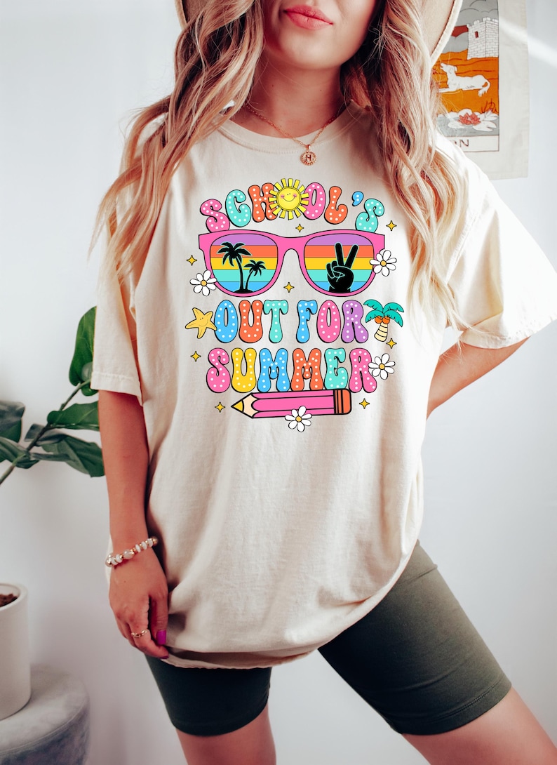 Op de afbeelding: Een wit T-shirt met een kleurrijk grafisch ontwerp met de tekst "School's Out For Summer". De grafiek toont een zonnebril met palmbomen en een vredesteken dat in de glazen wordt weerkaatst, een potlood en sterren en bloemen.