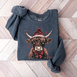 May include: A dark blue sweatshirt with a brown and white Highland cow wearing a red Santa hat and a red bow tie. The cow is surrounded by snowflakes.