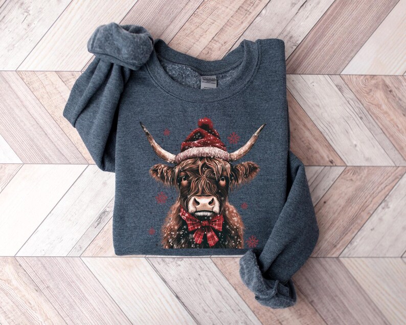 May include: A dark blue sweatshirt with a brown and white Highland cow wearing a red Santa hat and a red bow tie. The cow is surrounded by snowflakes.