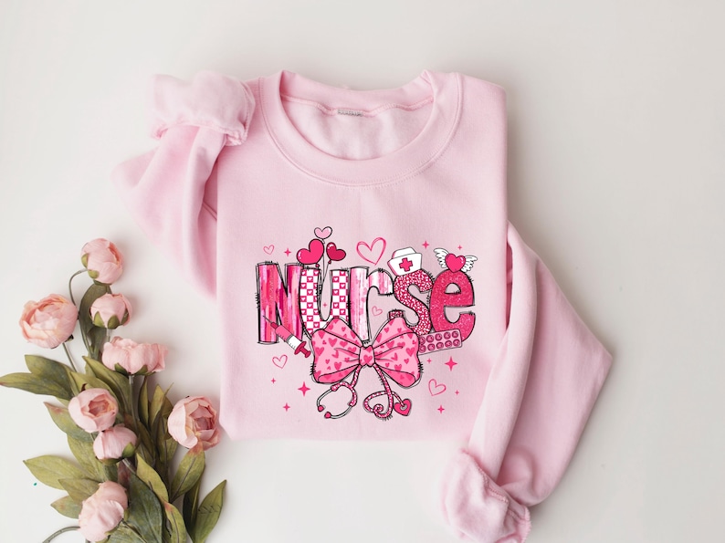 May include: Pink sweatshirt with a graphic design of the word "Nurse" in pink and white with hearts, a bow, a stethoscope, and other medical symbols.