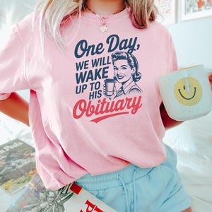 Puede incluir: Una camiseta rosa claro con el texto "One Day, We Will Wake Up To His Obituary" y una ilustraci&oacute;n vintage de una mujer sosteniendo una taza de caf&eacute;. La camiseta tambi&eacute;n presenta una taza blanca con una carita sonriente amarilla.