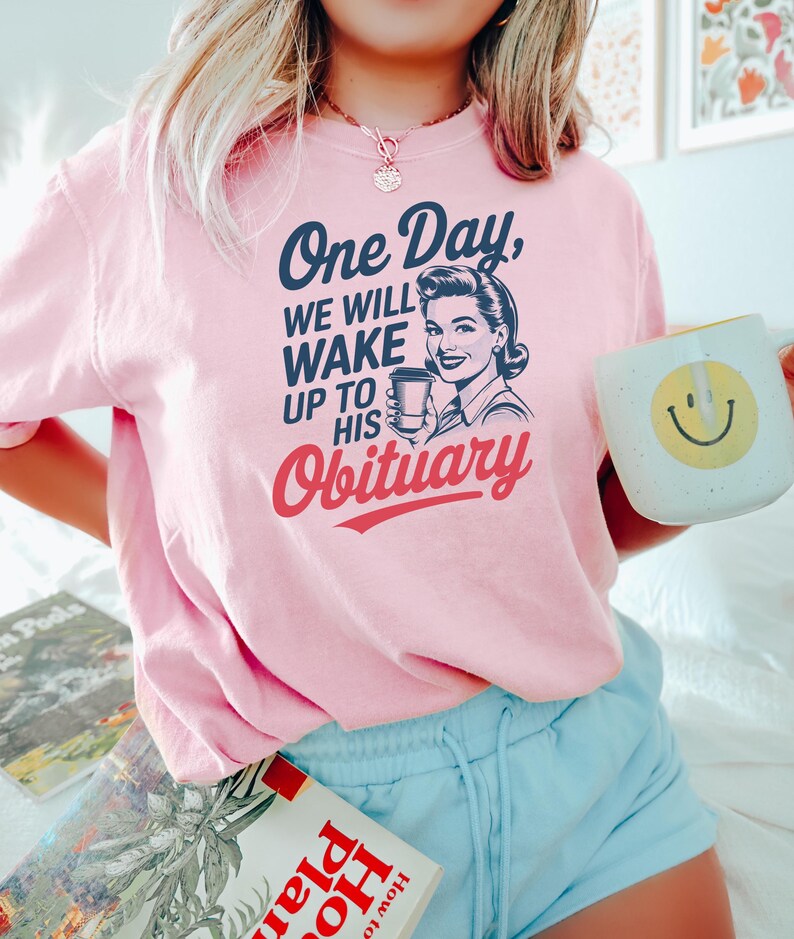 Puede incluir: Una camiseta rosa claro con el texto "One Day, We Will Wake Up To His Obituary" y una ilustraci&oacute;n vintage de una mujer sosteniendo una taza de caf&eacute;. La camiseta tambi&eacute;n presenta una taza blanca con una carita sonriente amarilla.