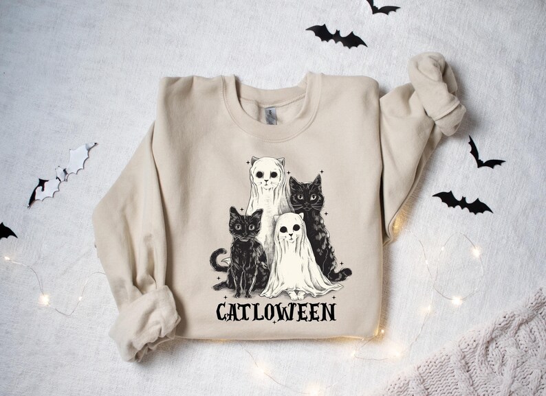May include: Beige sweatshirt featuring a graphic of four cats, two in black and two in white, with ghost-like coverings. The text "CATLOWEEN" is printed below the cats in a gothic font. Decorative bats are scattered around the sweatshirt.