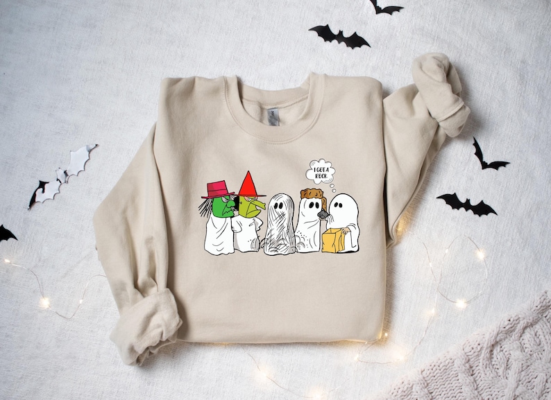 Puede incluir: Una sudadera beige con un estampado gr&aacute;fico de fantasmas y una bruja de dibujos animados. Los fantasmas llevan bolsas de caramelos y uno de ellos est&aacute; pensando "Tengo un libro".