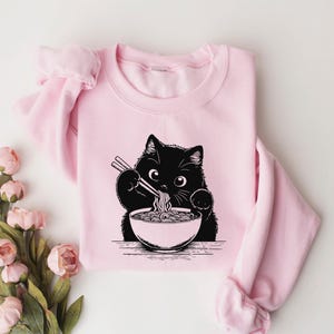 May include: Pink sweatshirt with a black cat eating ramen noodles. The cat is holding chopsticks and has a bowl of ramen in front of it.