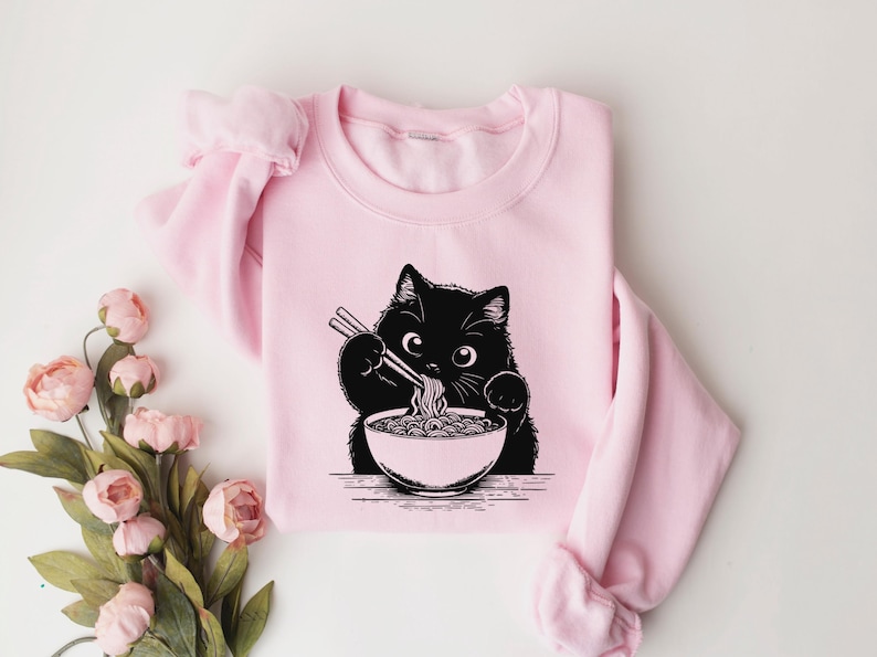 May include: Pink sweatshirt with a black cat eating ramen noodles. The cat is holding chopsticks and has a bowl of ramen in front of it.